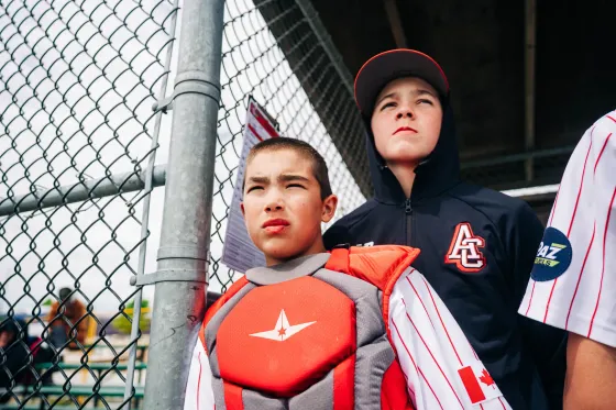 A young Abbotsford Cardinals catcher in full gear waits at the backstop fence.