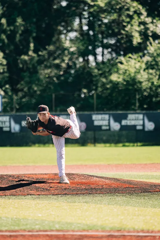 An Abbotsford Cardinals pitcher delivers with his leg high, a dark treeline behind him.