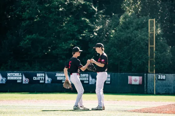Two Abbotsford Cardinals players meet on the mound, wide green field around them.