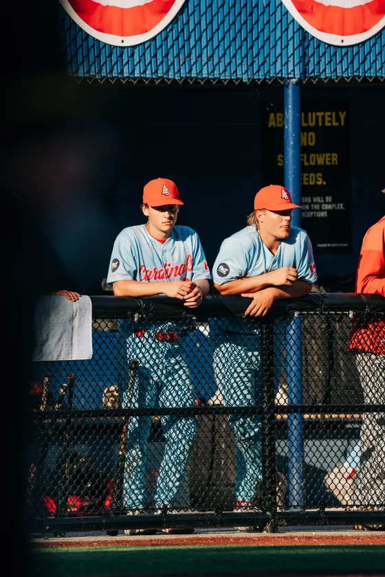 Two Abbotsford Cardinals players on the dugout rail, seen through the chain-link fence.