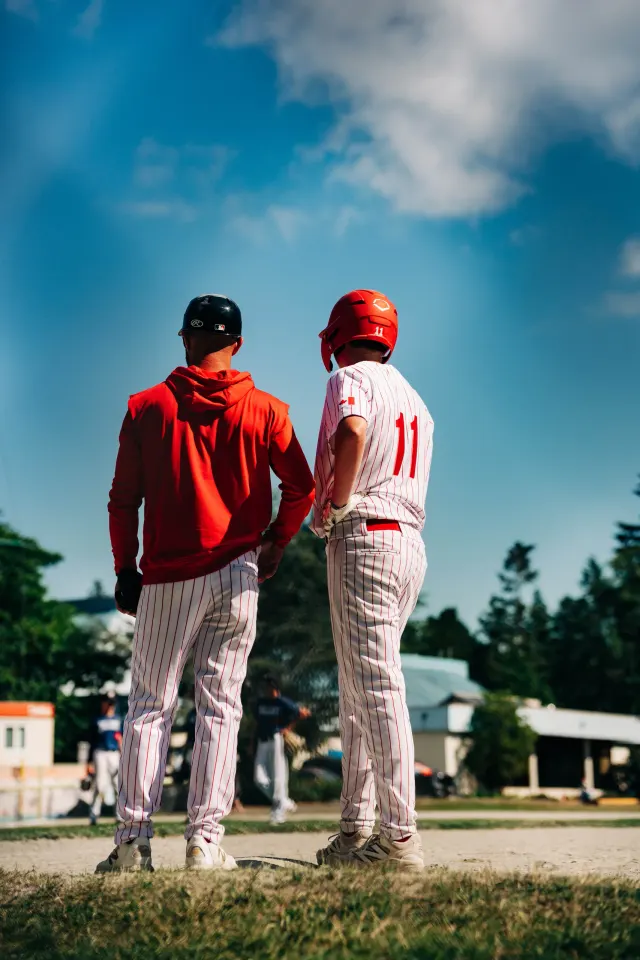 A Cardinals coach and a player in pinstripes stand together at the bag, both looking out across the field.