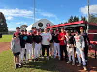A man in a white baseball sweatshirt stands in the middle of a group of Cardinals players in red and black at the dugout, smiling for a photo in front of the dome with the West Coast Baseball Collective and Abbotsford Cardinals badges.
