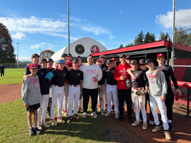 A man in a white baseball sweatshirt stands in the middle of a group of Cardinals players in red and black at the dugout, smiling for a photo in front of the dome with the West Coast Baseball Collective and Abbotsford Cardinals badges.